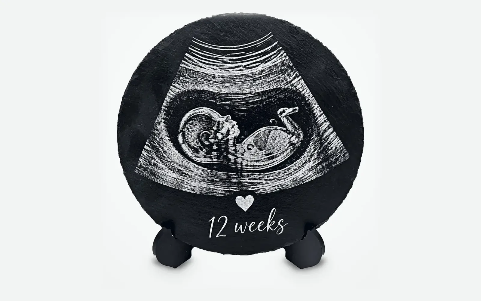 Round slate laser engraved and standing on its support, with a baby scan, a little heart and the words "12 weeks"